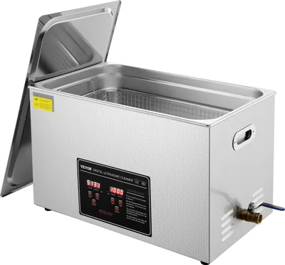 A silver ultrasonic cleaner with its lid open, revealing a stainless steel interior basket. The front panel features a digital display and controls, and a small drain valve is visible on the right side.