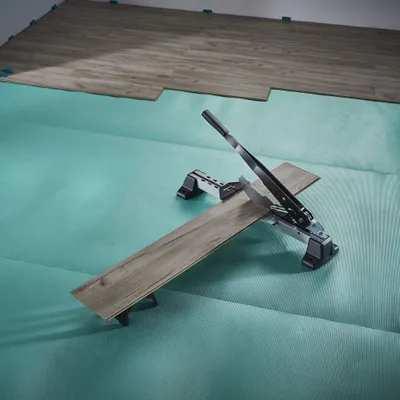 A vinyl and laminate cutter is being used to cut a plank of flooring on a teal underlayment.