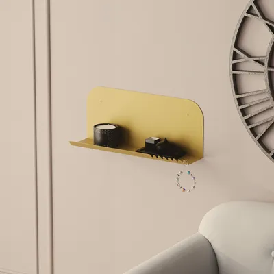 A gold shelf mounted on a light pink wall, displaying a black candle and a small black object, with a colorful bracelet hanging from the shelf. A large clock face is visible on the right.