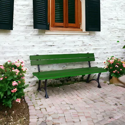 A green wooden garden bench sits on a brick patio in front of a white brick wall with a wooden window and black shutters. Pink flowers are in pots on either side of the bench.