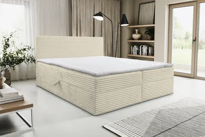 A beige and white striped boxspring bed with a mattress is shown in a well-lit room with curtains and a window in the background.