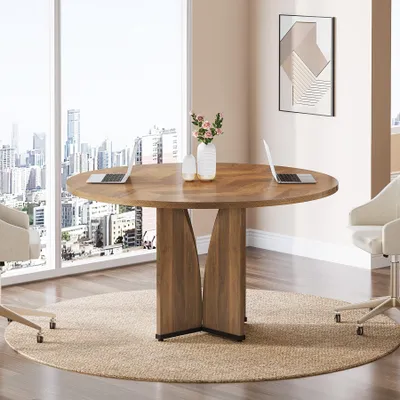 A round wooden conference table with laptops and vases in a modern office setting with a city view.