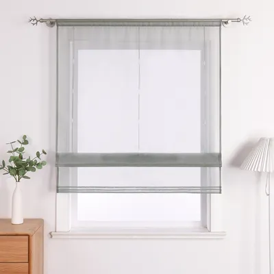 A sheer grey Roman blind is installed on a white window frame against a white wall. A small green plant in a white vase sits on a wooden bedside table to the left, and a white pleated lamp is visible to the right.