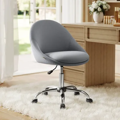 A gray fleece office chair with a silver base and wheels is placed on a white fluffy rug in front of a wooden vanity table.