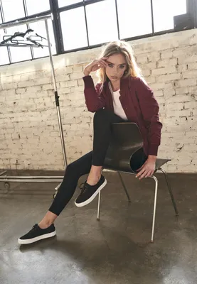 A woman is sitting on a chair in a studio setting with a brick wall and large windows in the background.