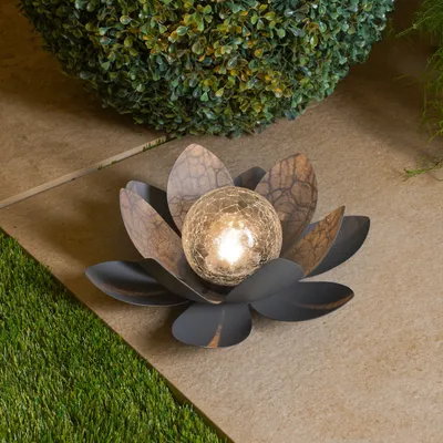A solar-powered lotus flower lamp with a cracked glass ball sits on a stone patio next to artificial grass and a bush.
