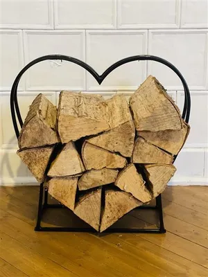 A heart-shaped metal log holder filled with firewood, placed in front of a white tiled wall and on a wooden floor.