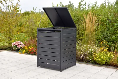 A dark gray garbage bin enclosure with its lid open, placed on a paved patio in a garden setting with various plants and flowers.