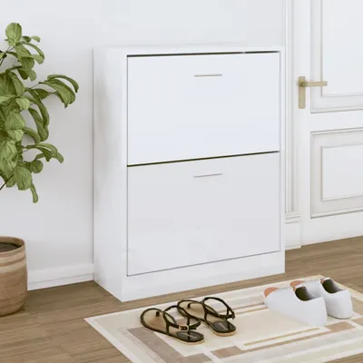 A white shoe cabinet with two drawers is shown in a room, with a plant to the left and a door to the right. Shoes are placed on a rug in front of the cabinet.