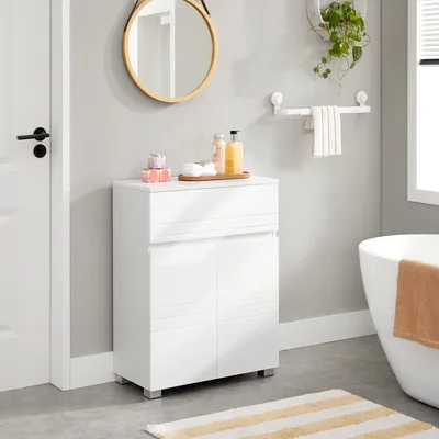 A white bathroom cabinet with a drawer and double doors, placed next to a white door in a bathroom setting. A round mirror hangs on the wall above the cabinet, and a bathtub is visible on the right.
