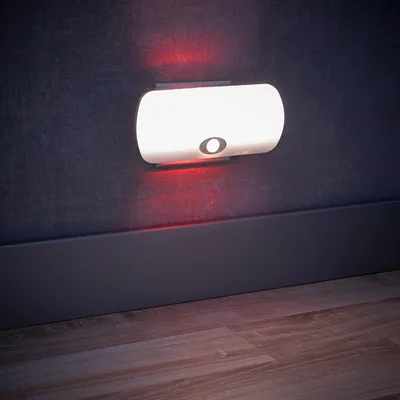 A white LED night light with a motion sensor is mounted on a dark textured wall above a dark gray baseboard. The floor in front of the baseboard is made of light brown wood planks.