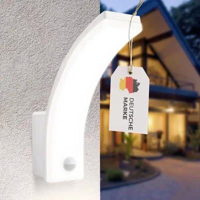 A white LED outdoor wall light with a motion sensor is mounted on a textured wall, with a tag indicating 'Deutsche Marke' (German Brand) hanging from it. The background shows a blurred image of a modern house at dusk.