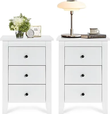 Two white nightstands are shown side-by-side against a white background. The left nightstand has a vase of flowers, a framed picture, and a clock on top. The right nightstand has a lamp, a book, and a cup and saucer on top. Both nightstands have three draw