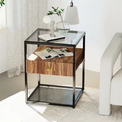 A modern side table with a glass top and a wood drawer is placed next to a white sofa, with a lamp and a vase of plants on top.