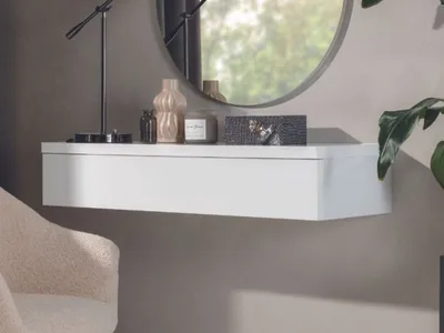 A white floating shelf mounted on a gray wall, decorated with various objects including a lamp, vase, candle, and a decorative box.