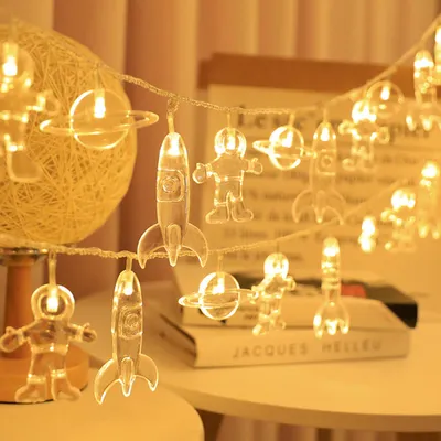 A string of decorative lights featuring planet, astronaut, and rocket shapes, illuminated with warm white light, displayed on a table with books in the background.