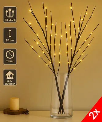 Two LED decorative branches with warm white lights in a glass vase, placed on a table with a candle.