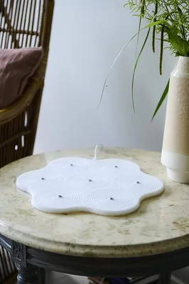 A white cloud-shaped object with circular indentations and small metal pins is placed on a marble-topped table next to a vase with greenery.
