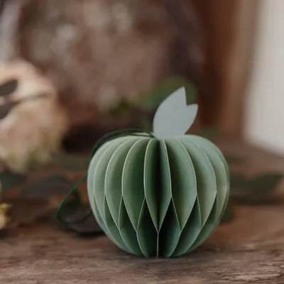 A green paper apple decoration sits on a wooden surface, with blurred foliage in the background.