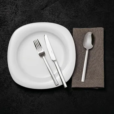 A white plate with a fork and knife, and a spoon on a brown napkin, set on a black textured surface.