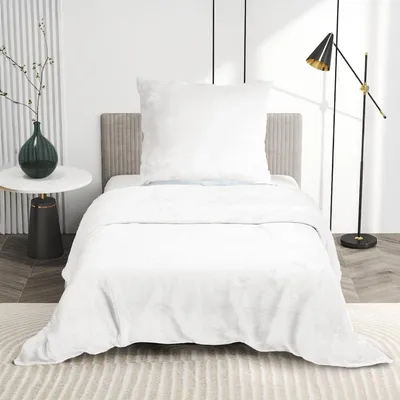 A white bed with white bedding and a large white pillow, set in a minimalist bedroom with a side table and a floor lamp.