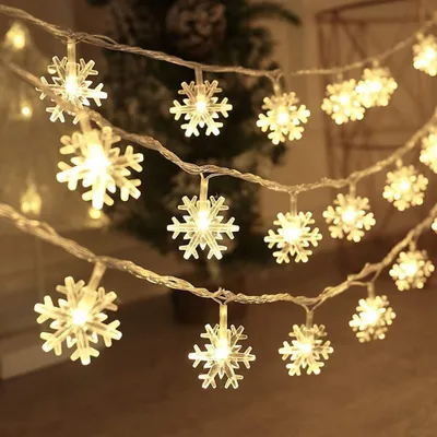A string of snowflake-shaped lights is draped across a dark background, with a blurred Christmas tree visible behind it.