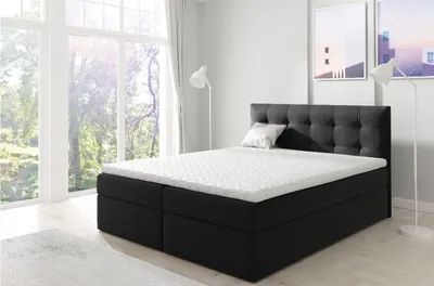 A black upholstered boxspring bed with a white mattress and topper is shown in a bright, modern bedroom with large windows.