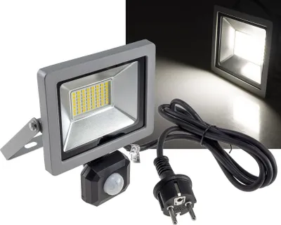 A floodlight with a motion sensor and a power plug is shown against a dark background, with a second floodlight illuminated in the background.