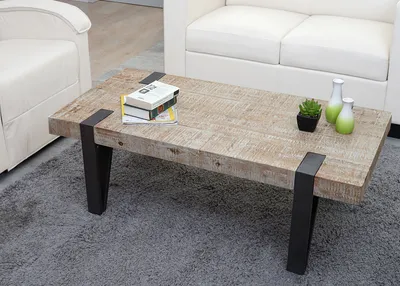 A rustic wooden coffee table with black metal legs, placed on a gray rug in a living room setting with white sofas in the background.