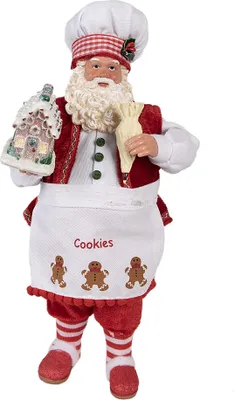 A Santa Claus figurine dressed as a baker, holding a gingerbread house and a piping bag.