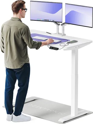 A man stands at a white standing desk with two monitors, using a treadmill mat on the floor.