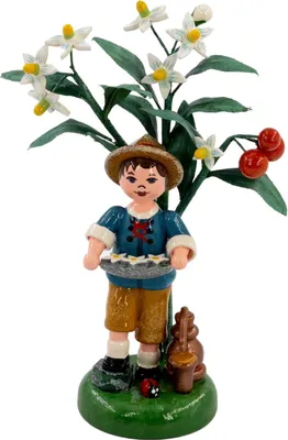 Ein kleiner Junge in traditioneller Kleidung hält ein Tablett mit weißen Blüten und steht vor einem Hintergrund aus grünen Blättern und weißen Blüten mit roten Kirschen.