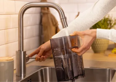 A person is rinsing a coffee maker's water tank under a faucet in a kitchen sink.