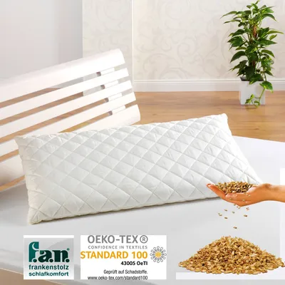 A white quilted pillow is shown on a white surface, with a wooden headboard behind it. A hand is pouring grain into a pile next to the pillow. There are also certification logos at the bottom.