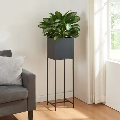 A black metal plant stand with a green plant in a black pot, placed next to a gray sofa and a window with white curtains.
