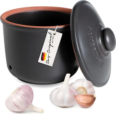 A black ceramic garlic pot with a lid, alongside three garlic bulbs. A tag hangs from the pot.