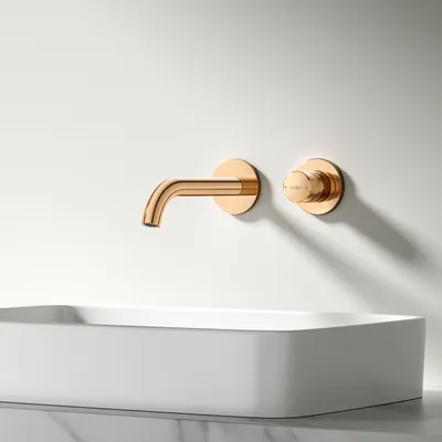 A rose gold faucet and control knob are mounted on a white wall above a white rectangular sink.