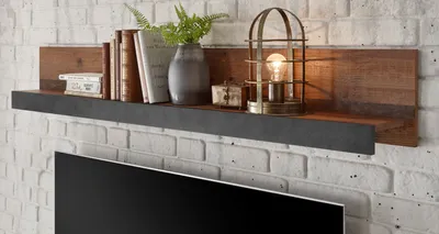 A wooden shelf with books, a vase, and a lamp mounted on a white brick wall above a television.