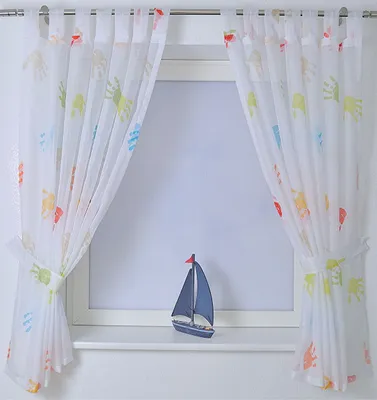 A pair of sheer curtains with colorful handprints hanging in front of a window, with a small model sailboat on the windowsill.