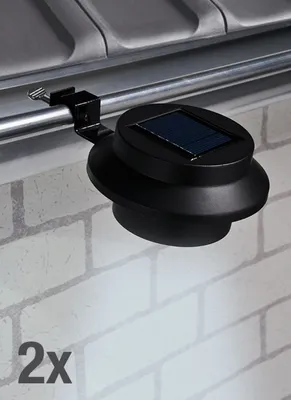 A black solar-powered gutter light is clamped onto a metal railing, with a brick wall in the background.