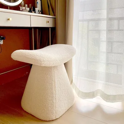 A fluffy white stool sits in front of a vanity table near a window with sheer curtains.