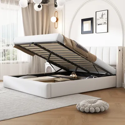 A white upholstered bed frame with the mattress lifted to reveal storage space underneath. The bed is in a room with curtains and wall art.