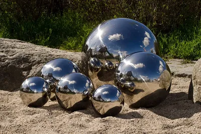 Several polished stainless steel spheres of varying sizes are arranged on a sandy surface next to large rocks, with greenery in the background.