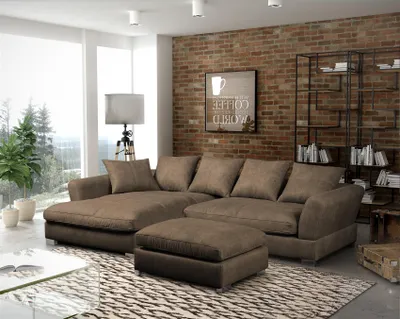 A modern living room with a large sectional sofa and ottoman, set against a brick wall and large windows.