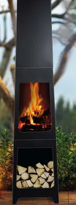 A tall, black outdoor fireplace with a fire burning inside and firewood stored below, set against a blurred background of trees and greenery.
