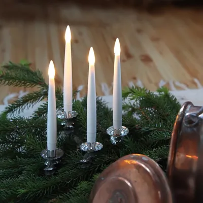 Four LED taper candles with flickering flames are arranged on a bed of evergreen branches, with copper pots in the foreground.