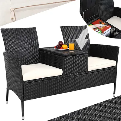 A black rattan garden bench with white cushions and a central table. The table has a glass of orange juice and fruit on it. An inset shows the storage compartment of the table open with books inside.