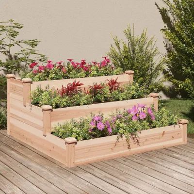 A tiered wooden planter box filled with flowers and plants on a wooden deck, with a wall and greenery in the background.