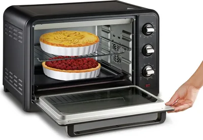 A hand is shown opening the door of a black electric oven. Inside the oven, two pies are visible on racks.