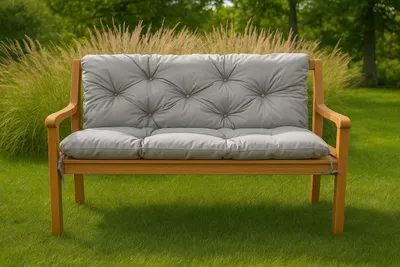 A garden bench with a gray cushion sits on a green lawn with tall grasses and trees in the background.